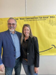A man wearing glasses and a suit smiling next to a woman smiling and wearing a suit, in front of a large yellow back drop that reads 'The Make Construction Fun Again Wall'