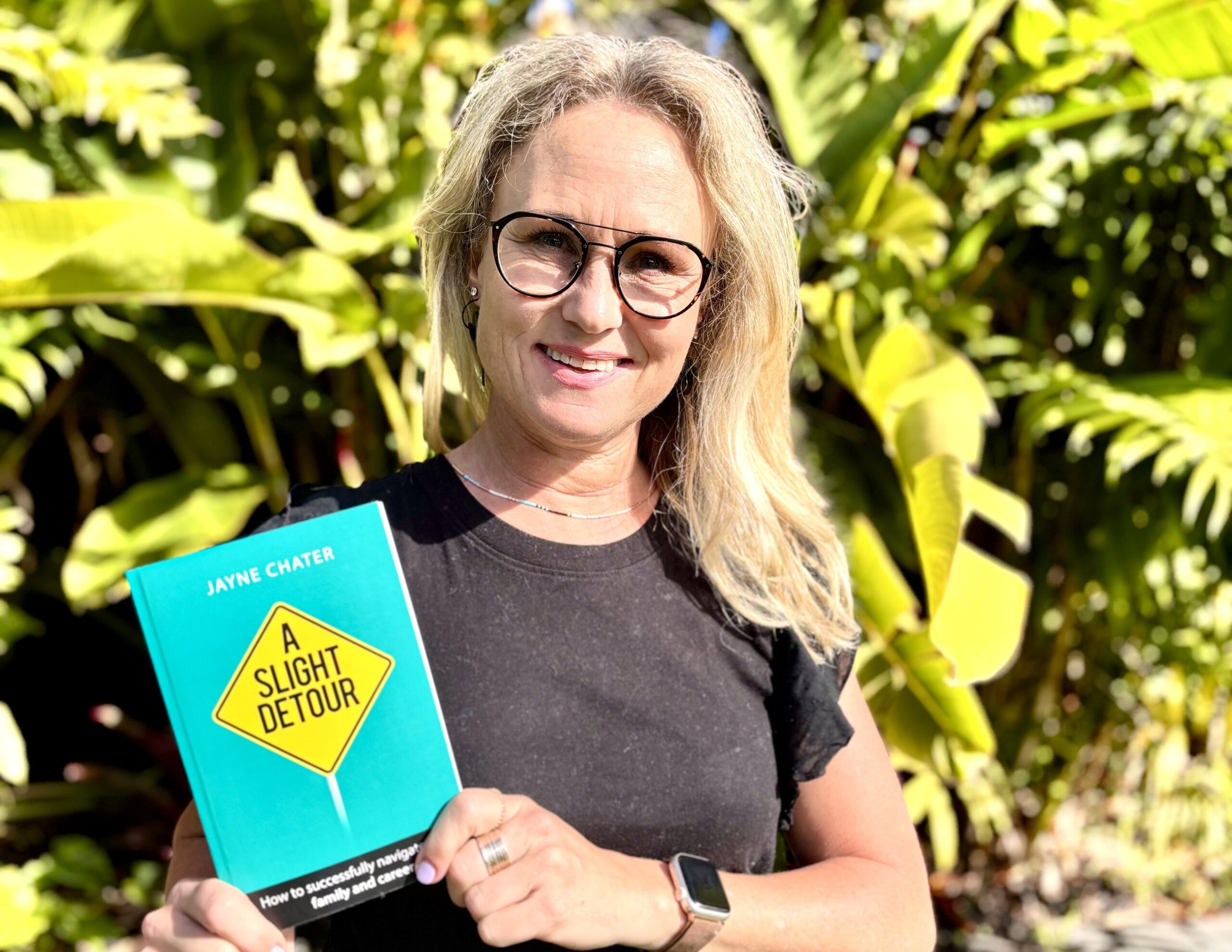 Jayne Chater, a smiling blonde woman wearing glasses, holds her book A Slight Detour